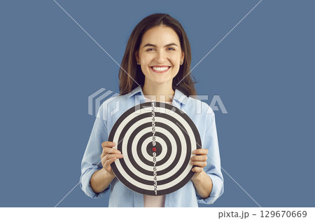 Positive smiling brunette woman in casual shirt holding darts shooting target on blue background. 129670669