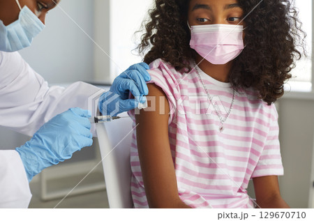 African american doctor giving a vaccine injection vaccination to teen girl in shoulder in clinic. 129670710