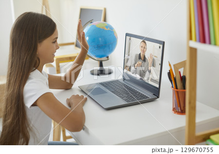 Happy school girl learning online, having remote class and waving hand at laptop screen 129670755
