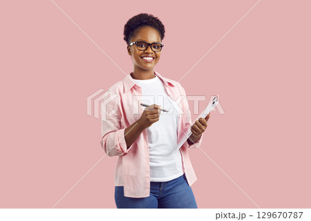 Happy smiling African American woman with pen and clipboard standing on pink background 129670787