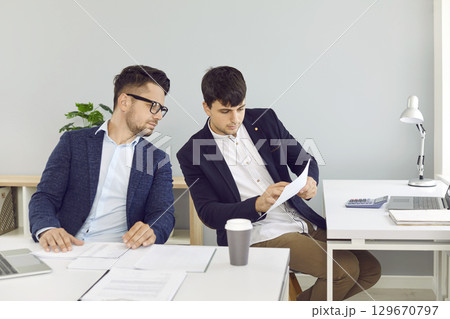 Two male employees sitting at office desks and looking at some business documents Two male employees sitting at office desks and looking at some business documents 129670797