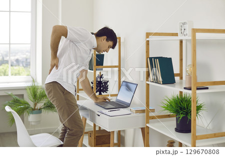 Man who works at laptop feels pain in his back, gets up and massages his back to relax. 129670808