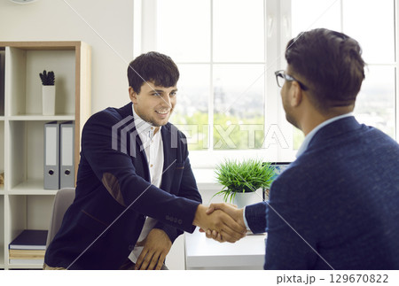 Two people sitting at an office desk, making a business deal and exchanging handshakes Two people sitting at an office desk, making a business deal and exchanging handshakes 129670822