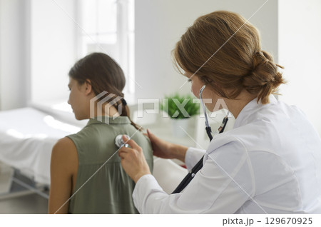 Girl in clinic for a checkup. Doctor with stethoscope examining child's lungs and heartbeat. 129670925