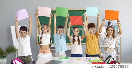 Happy elementary school boys and girls hold up colored paper sheets in classroom. Happy elementary school boys and girls hold up colored paper sheets in classroom. 129670928