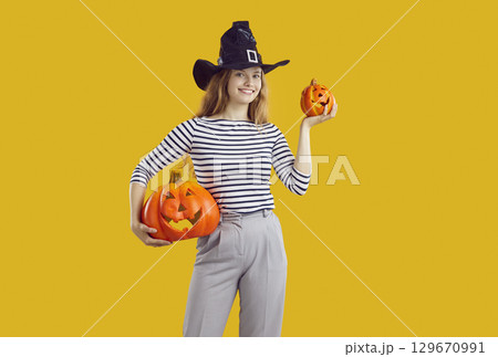 Young attractive student-age woman holds two Halloween pumpkins stands on orange studio background 129670991