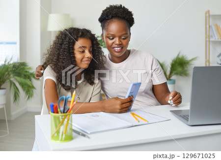 Happy school child and mother sitting at desk, doing homework and using mobile phone 129671025