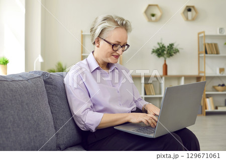 Mature woman in glasses is working typing laptop sitting on couch in her cozy office. 129671061