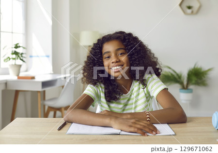 Smiling african american teen girl is studying online looking at camera sitting at desk at home. 129671062