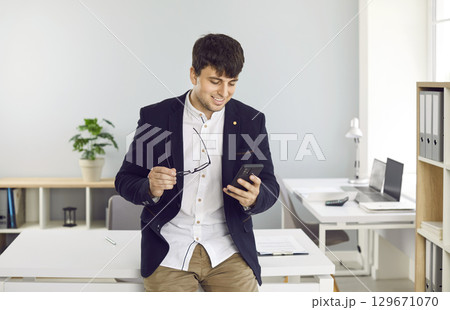 Satisfied young businessman using mobile phone by phone near workplace in his office. Satisfied young businessman using mobile phone by phone near workplace in his office. 129671070