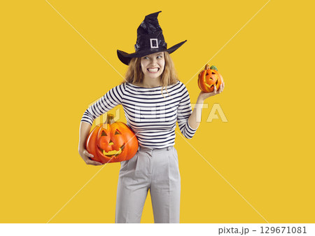 Young woman in witch hat holding Halloween pumpkins and making funny, scary face 129671081