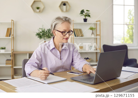 Senior business lady is comparing electronic and paper documents while working in her home office. Senior business lady is comparing electronic and paper documents while working in her home office. 129671155