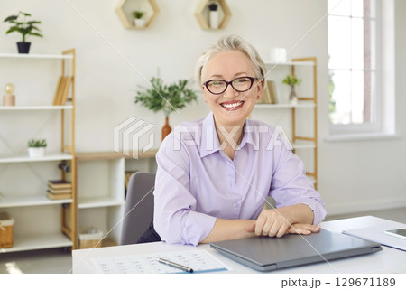 Gray haired elderly Caucasian woman smiling looking at camera sits at home office desk with laptop 129671189