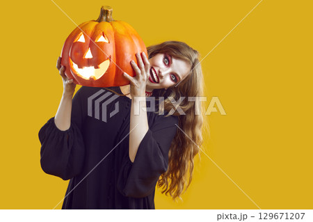 Woman with scary make-up dressed in black gown is holding Halloween pumkin on yellow background. Woman with scary make-up dressed in black gown is holding Halloween pumkin on yellow background. 129671207