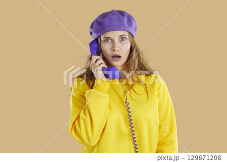 Stunned puzzled woman in purple hat and yellow sweatshirt calling phone on beige background. Stunned puzzled woman in purple hat and yellow sweatshirt calling phone on beige background. 129671208