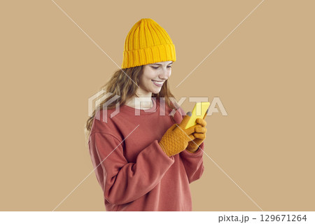 Happy smiling woman in yellow hat and gloves is looking at smartphone on beige background. 129671264