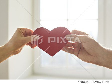 Close up of young couple holding red paper heart in hands against window background Close up of young couple holding red paper heart in hands against window background 129671266