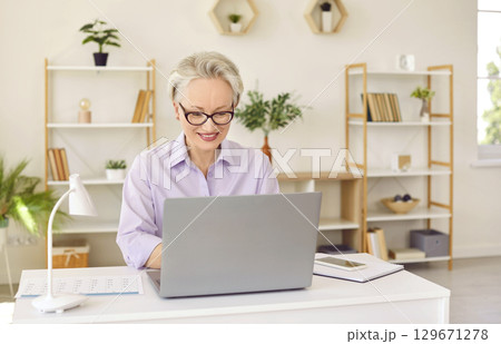 Senior positive smiling woman in glasses and blouse wirking on laptop sitting at desk in office. Senior positive smiling woman in glasses and blouse wirking on laptop sitting at desk in office. 129671278