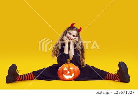 Happy woman in Halloween costume sitting on yellow studio background with Jack o lantern 129671327