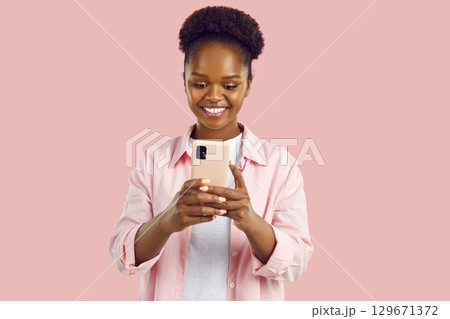 Happy smiling african american woman is looking at smartphone screen on light pink background. Happy smiling african american woman is looking at smartphone screen on light pink background. 129671372