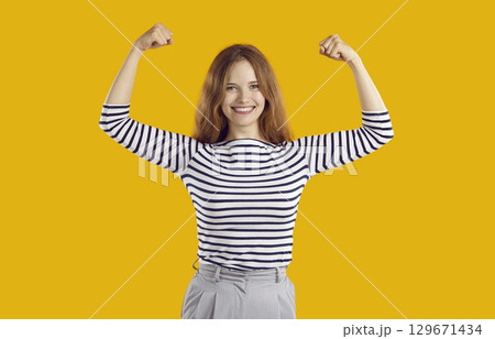 Happy confident beautiful young woman smiling, flexing her arms and showing girl power Happy confident beautiful young woman smiling, flexing her arms and showing girl power 129671434