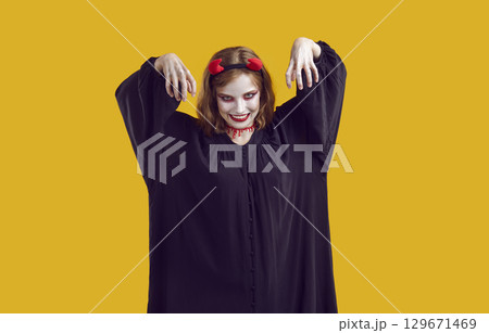 Woman in black robe, makeup, devil horns, bloody trail on her neck scares audience with her hands up. Woman in black robe, makeup, devil horns, bloody trail on her neck scares audience with her hands up. 129671469