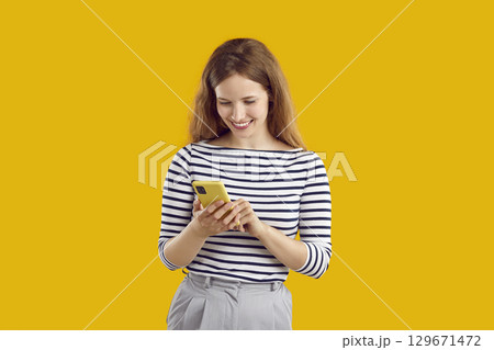 Happy smiling woman in striped sweatshirt is browsing her smartphone on yellow background. 129671472