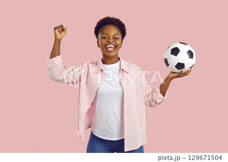Soccer fan woman saying yes showing win gesture holding football ball in hand on pink background. Soccer fan woman saying yes showing win gesture holding football ball in hand on pink background. 129671504