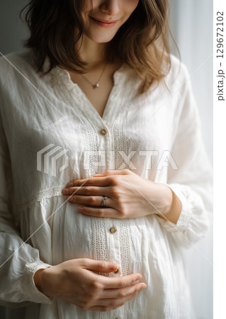 白い服を着た妊婦が優しくお腹を抱える女性。マタニティポートレート 129672102