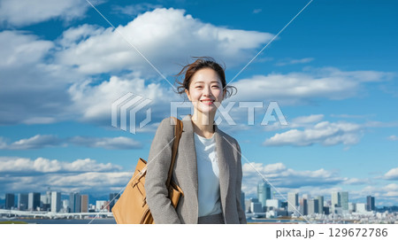 青空の下で笑顔を見せるビジネスウーマンと都市風景。日本人の若い女性のライフスタイル 129672786