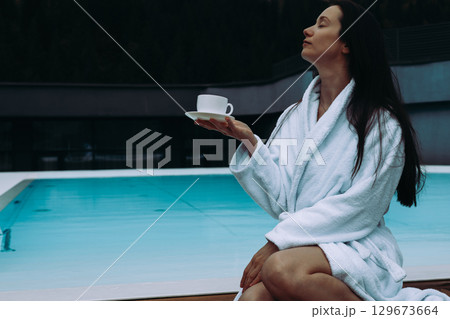 A relaxed woman enjoys a cup of tea near a pool, wearing a white robe. A serene lady in a white robe enjoys a moment with a cup of tea in spa hotel . 129673664
