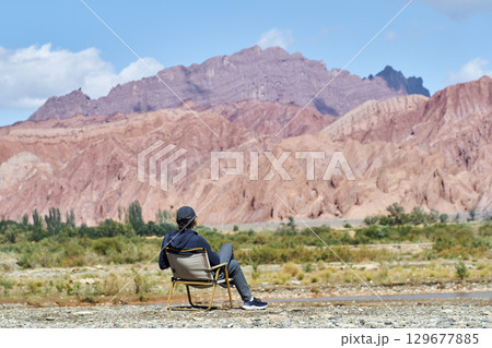 asian man sitting in a chair looking at a reddish mountain 129677885