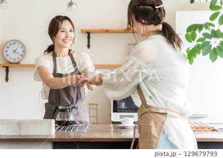 キッチン・カウンターからコーヒーカップを手渡すエプロン姿のカフェ店員のアジア人女性 129679793