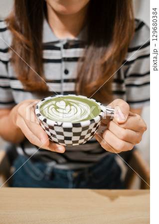 Close-up of a woman holding a matcha latte with a heart design, wearing a striped shirt in a cozy cafe. 129680384