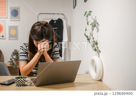 A woman sits at a desk with a laptop, appearing stressed, in a modern home office environment. 129680400