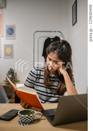 A young woman reads a book at her home office desk, surrounded by a laptop and smartphone, creating a cozy and modern workspace atmosphere. 129680406