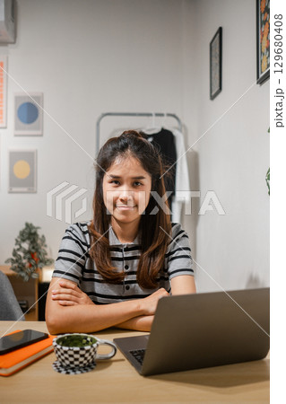 Young woman sitting at a desk with a laptop and coffee, exuding confidence in a modern home office setting. Young woman sitting at a desk with a laptop and coffee, exuding confidence in a modern home office setting. 129680408