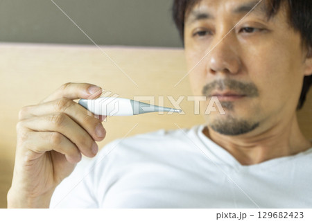 Sick man with fever laying in bed taking temperature with thermometer 129682423
