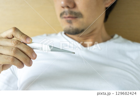 Sick man with fever laying in bed taking temperature with thermometer Sick man with fever laying in bed taking temperature with thermometer 129682424