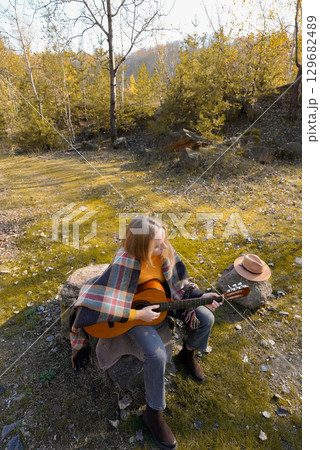 Beautiful blonde girl playing acoustic guitar and singing relaxing enjoying outdoors on sunny autumn day. Hippie woman in boho style learning and practicing playing musical instrument. Fall leaves 129682489