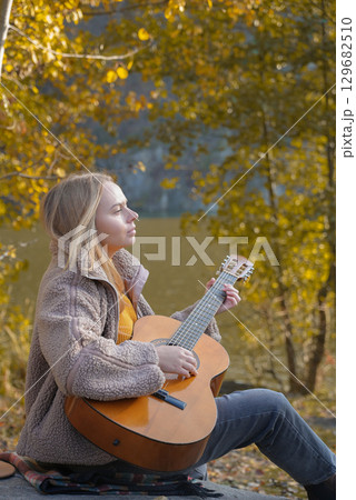 Beautiful blonde girl playing acoustic guitar and singing relaxing enjoying outdoors on sunny autumn day. Hippie woman in boho style learning and practicing playing musical instrument. Fall leaves Beautiful blonde girl playing acoustic guitar and singing relaxing enjoying outdoors on sunny autumn day. Hippie woman in boho style learning and practicing playing musical instrument. Fall leaves 129682510