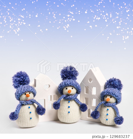 Knitted snowman in blue hat and scarf with white houses on gray background. Winter fairy tale story with your own hands, hobby. Merry Christmas and New Year 2025 greeting card 129683237