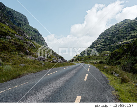 The road ahead, an inviting asphalt ribbon, promises a journey through dramatic Irish countryside. Towering green hills flank the path to untold adventures. The road ahead, an inviting asphalt ribbon, promises a journey through dramatic Irish countryside. Towering green hills flank the path to untold adventures. 129686157