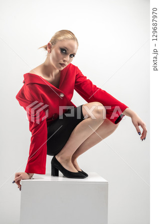 Beautiful woman with red jacket and mini shirt sitting on white cube 129687970