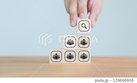 Hand placing a magnifying glass icon wooden cube block above people group wood blocks, representing talent search, company, leadership, recruitment, or leadership selection strategy. 129690930