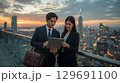 夕暮れの都市高層ビル屋上でタブレットを見ながら商談する日本人ビジネスパーソン 129691100