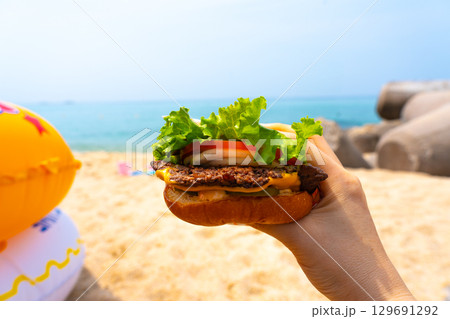 Woman holding burger in a hand against beach background 129691292