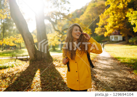 Happy female hiker in yellow coat walking in golden autumn forest. Woman has fun in autumn in park. 129694104