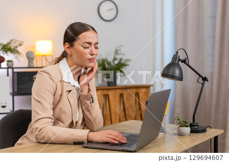 Young businesswoman touches cheek feels tooth pain while working on laptop and slows typing pace Young businesswoman touches cheek feels tooth pain while working on laptop and slows typing pace 129694607
