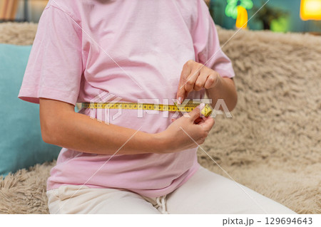 Pregnant woman measuring belly with tape smiling satisfied with progress expecting baby birth soon 129694643